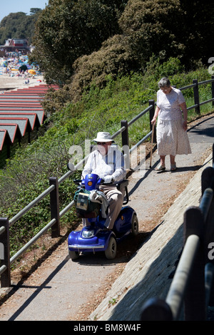 Il vecchio gentleman nel cappello per il sole sui disabili motorizzata buggy seguita da una vasta con bastone scendendo la rampa verso la spiaggia Foto Stock