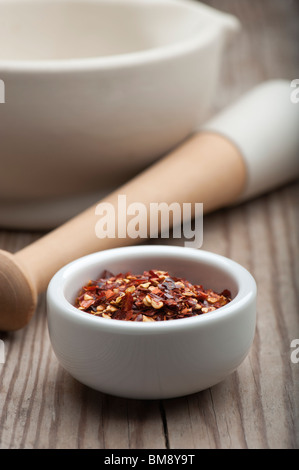 Peperoncino frantumato in un piatto bianco, su di un tavolo di legno con un mortaio e pestello in background Foto Stock