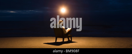 Uomo seduto nella sedia sotto la lampadina di luce sulla spiaggia di notte, libro di lettura Foto Stock