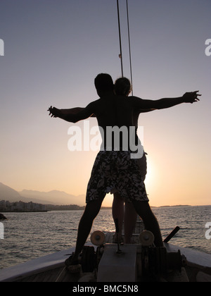 KYRENIA, CIPRO DEL NORD. Una giovane coppia in piedi sulla prua di un caicco turco e barca a vela al tramonto. Foto Stock