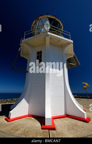 Vista verso l'alto di un faro bianco con base rossa, situato vicino a un oceano, e segnali direzionali visibili. Cape Reinga, nuova Zelanda Foto Stock