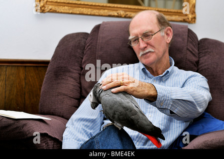 A 70 anni di vecchio uomo caucasico si siede in una poltrona reclinabile petting il suo animale domestico pappagallo grigio africano. Foto Stock