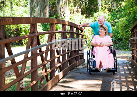 Senior uomo spinge la sua moglie di disabili su sedia a rotelle del attraverso il parco. Foto Stock