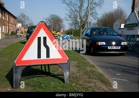 Auto passando un cartello stradale e barriere su una strada inglese durante i lavori di riparazione. Foto Stock