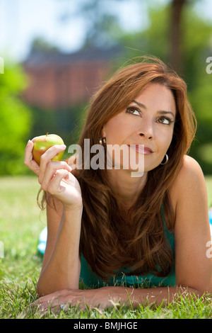 Una bella donna nella sua anni trenta che stabilisce fuori sull'erba azienda o mangiare un apple cercando e di pensiero Foto Stock