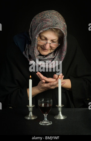 Un'anziana donna ebrea, testa coperta con una sciarpa, prega oltre due accesa sabbath candele e vino di sabato Foto Stock