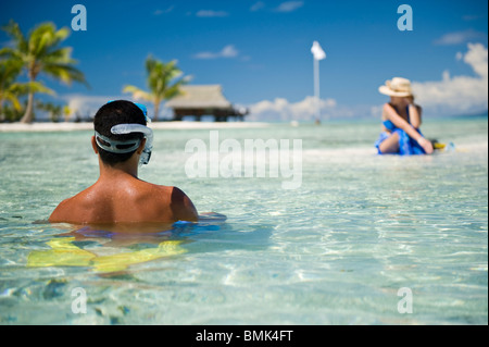 Un uomo e una donna seduta in acque poco profonde su una spiaggia di sabbia bianca. La donna è prendere il sole, l'uomo è di andare a fare snorkeling Foto Stock