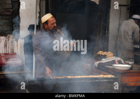 Fornitore di cibo fanning il fumo come egli cuochi nei vicoli di Kashgar nella Western Xinjiang provincia di PR Cina. Foto Stock