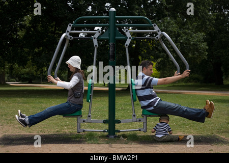 Due genitori provare macchine ginniche in un parco pubblico come bambino gioca al di sotto. Foto Stock