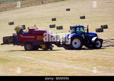 Fattoria di blu il trattore e la rotopressa di fieno fieno la raccolta su andane, agricoltura in sunny Wales UK. Il lato orizzontale vista. Foto Stock