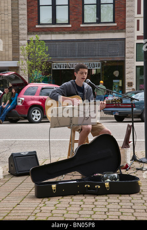 Un giovane artista di strada che suona su una chitarra acustica per la strada della città con cassa aperta per chitarra e denaro in formato verticale americano ad alta risoluzione Foto Stock
