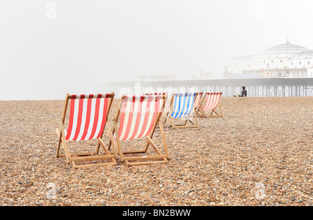 Blu e rosso di sedie a sdraio su una nebbia spiaggia ghiaiosa con il molo di Brighton in background Foto Stock