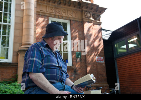 Un uomo seduto su una parete la lettura di un libro al di fuori di una biblioteca pubblica, Loughborough, Leicestershire, Inghilterra. Foto Stock