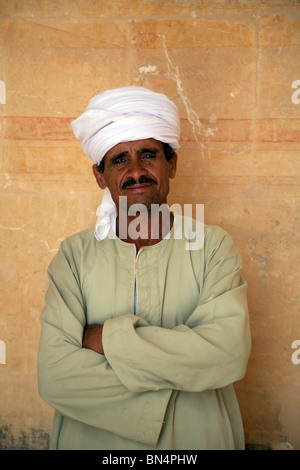 Un uomo egiziano indossando un tradizionale galabia stare vicino a un muro, il tempio mortuario di Hatshepsut, Luxor, l'Alto Egitto Foto Stock