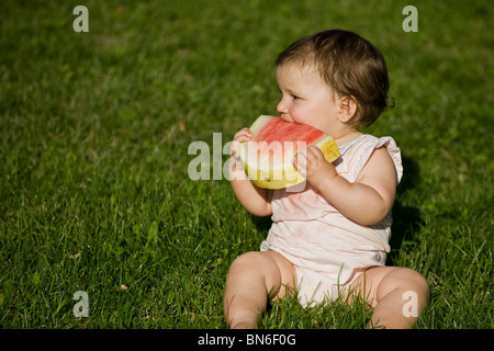 Un simpatico bambina bambino neonato Bambino, si siede in un campo erboso godendo di un rinfrescante anguria durante una calda giornata estiva Foto Stock