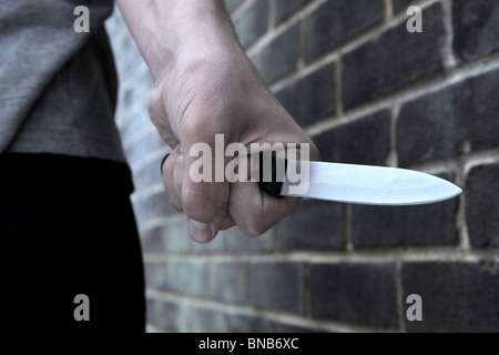 Canto di un giovane maschio in possesso di un coltello camminare davanti a un muro di mattoni Foto Stock