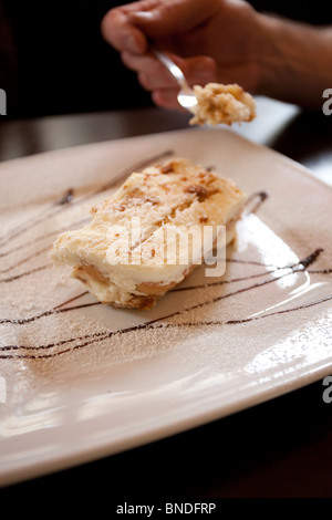 Una deliziosa fetta di tiramisù su un piatto quadrato bianco, spolverato di cacao e adornato con salsa al cioccolato, accompagnato da una mano che tiene una forchetta Foto Stock