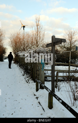 L'uomo, avvolto contro il freddo, salire a piedi a percorso nella neve con turbina eolica in distanza Gillespie il Parco di natura di Highbury London REGNO UNITO Foto Stock