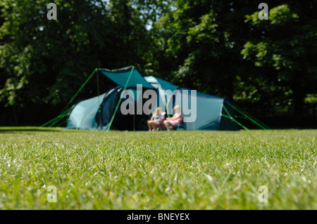 Due giovani ragazze sat al di fuori di una grande famiglia di verde tenda inondata di sole in un campeggio con alberi dietro Foto Stock