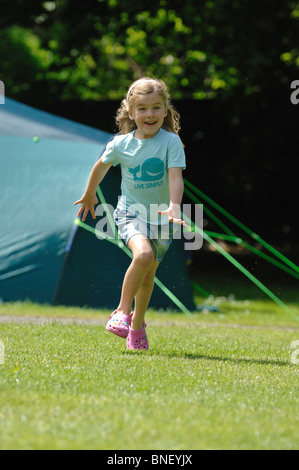 Una giovane ragazza in un turchese T-shirt a correre verso la telecamera e ridere con una verde grande famiglia tenda in background. Foto Stock