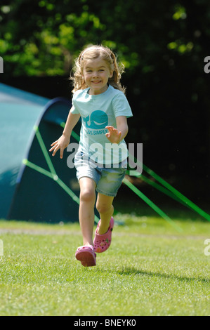 Una giovane ragazza in un turchese T-shirt a correre verso la telecamera e ridere con una verde grande famiglia tenda in background. Foto Stock