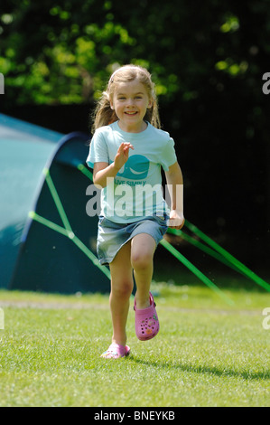 Una giovane ragazza in un turchese T-shirt a correre verso la telecamera e ridere con una verde grande famiglia tenda in background. Foto Stock