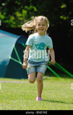 Una giovane ragazza in un turchese T-shirt a correre verso la telecamera e ridere con una verde grande famiglia tenda in background. Foto Stock