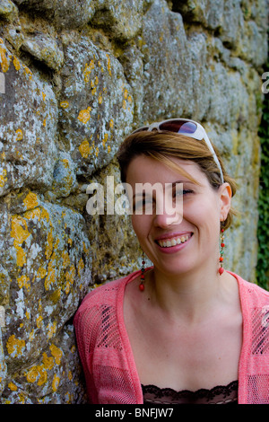 Sorridente attraente dama appoggiata contro un vecchio usurato meteo pietra costruito il muro. Lei è nei suoi primi anni trenta. Foto Stock