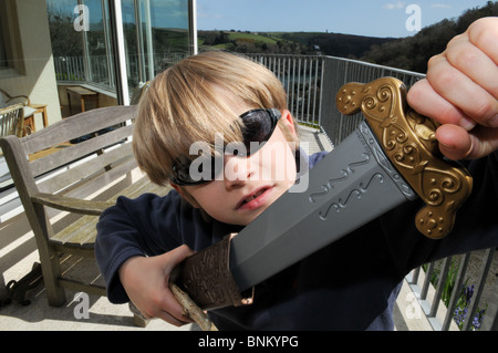 Biondo ragazzo che indossa gli occhiali da sole e giocare con una spada di plastica e fodero esterno da un banco di lavoro. Foto Stock