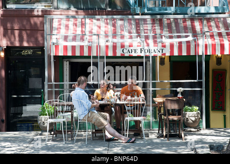 Animated giovane donna con 2 amici maschi rilassante a tree ombreggiati tavoli esterni posti a sedere del piccolo e affascinante caffetteria sulla giornata calda NYC Foto Stock