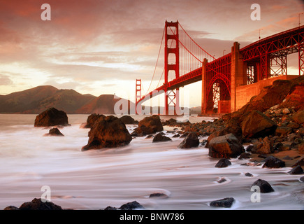 Forme d'onda colpendo costa rocciosa lungo Baker Beach sotto il Ponte Golden Gate a San Francisco al tramonto Foto Stock