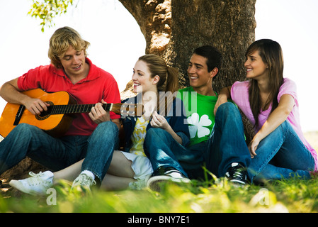 Giovane uomo seduto con gli amici sotto agli alberi, suonare la chitarra Foto Stock