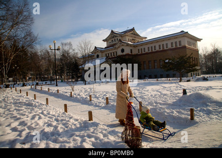 La Russia, Estremo Oriente, Sakhalin Yuzhno-Sakhalinsk;; una giovane donna spingendo una slitta da neve con il suo bambino di fronte ad uno dei musei Foto Stock