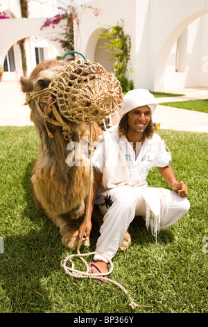 Un uomo si siede accanto a un cammello dromedario presso un hotel sull'isola di Djerba, Tunisia. Foto Stock