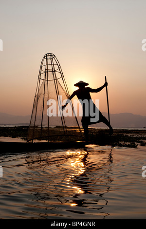 Una gamba di canottaggio manovre pescatore il suo legno teak canoa sopra il pesce sul Lago Inle come il sole tramonta dietro di lui che delinea il suo corpo un Foto Stock