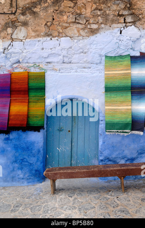 La visualizzazione di tessuti colorati tappeti tappeti sospesi o tappeti per la vendita, blu porta e panca in legno, Chefchaouen, Marocco Foto Stock