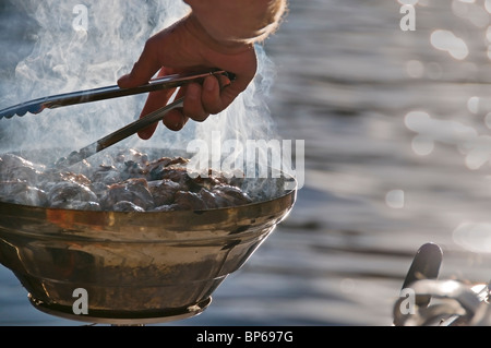 Un uomo trasforma i pezzi di pollo con pinze su un caldo barbecue grill attaccato alla parte posteriore di una barca in acqua di Puget Sound. Foto Stock