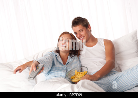 Giovane uomo e donna sdraiata nel letto bianco a guardare la televisione e mangiare patatine Foto Stock