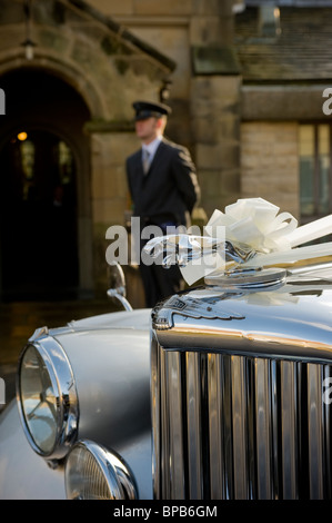 Primo piano di un'auto Jaguar decorata con nastri bianchi per un matrimonio, con autista fuori fuoco in piedi fuori da una chiesa, in lontananza. Foto Stock
