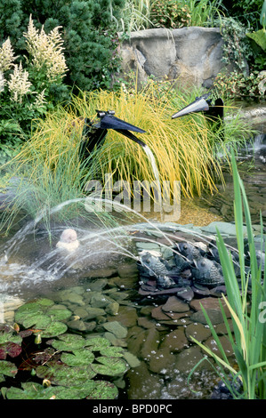 L'acqua da giardino con buffo uccello fontane, rane schizzando torrenti nel laghetto in giardino con piante lilypads Graminacee ornamentali Foto Stock