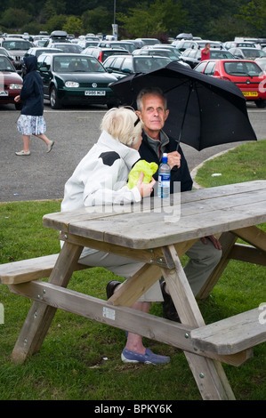 L uomo e la donna seduta su una panchina nel parco vicino ad un parcheggio sotto la pioggia sotto un ombrello. Looe, Cornwall, Regno Unito Foto Stock