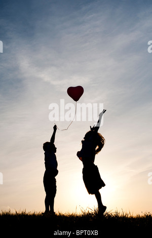 Silhouette di un giovane ragazzo e ragazza che gioca con il cuore di un palloncino di forma al tramonto Foto Stock