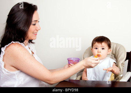 Bella madre felice o nanny alimenta funny baby boy girl purea di arancia con cucchiaio, neonati mangia confuso, seduti a tavola. Foto Stock