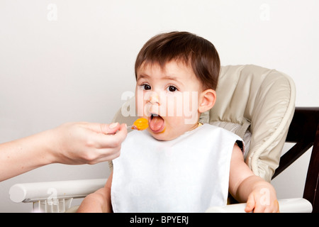 Bella happy baby infant boy ragazza con la bocca aperta mangiare confuso purea di arancia dal cucchiaio. Foto Stock