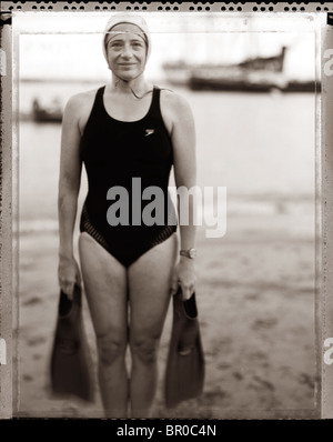 Di mezza età donna caucasica nuotatore su una spiaggia indossando un oscuro costume da bagno azienda pinne in ogni mano. Foto Stock
