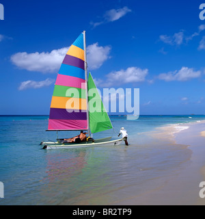 Uomo che lancia barca a vela colorata catamarano da bordo acque di spiaggia tropicale coppia vacanza a bordo a Tamarind Cove Barbados Caraibi Foto Stock