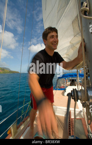 Un uomo hoistes vela principale a bordo di una barca a vela, Hanalei Bay, Costa di Na Pali, Kauai, Hawaii (movimento sfocata) Foto Stock