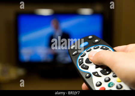 Mano che tiene il telecomando del TV con una televisione in background. Close up. Foto Stock
