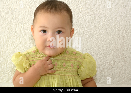 Tulum, Quintana Roo/Messico - 4 maggio: Baby girl in abito verde cercando per la fotocamera con la mano sul mento posti a sedere contro il muro bianco Foto Stock