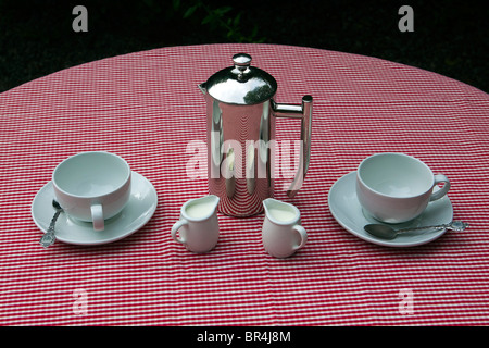 A shiny coffee pot and cups and saucers on a red and white tablecloth Foto Stock